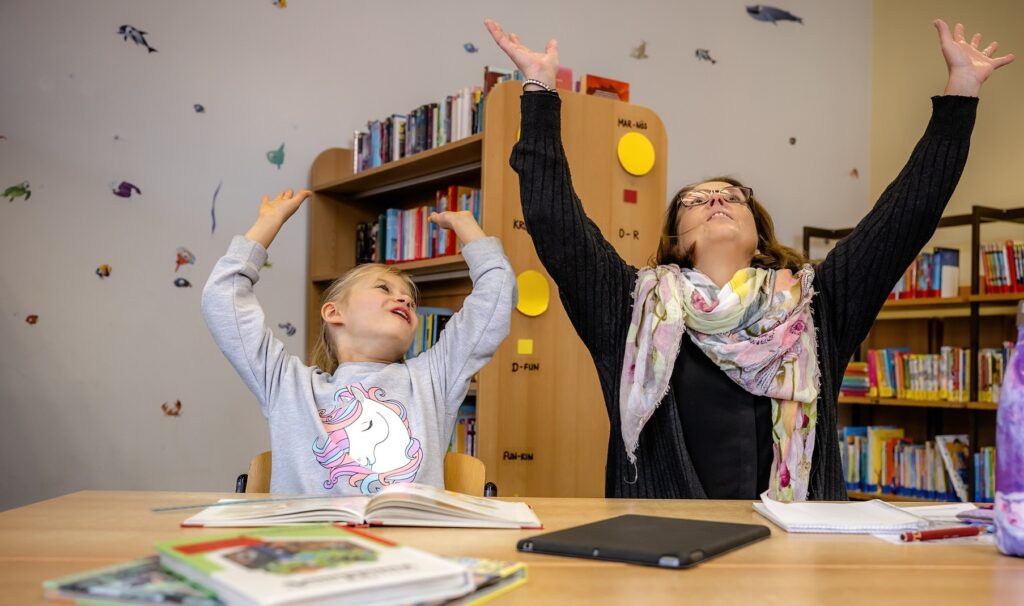 Foto von einem Mädchen und einer Frau, die gemseinsam an einem Tisch sitzen und beide Arme in die Luft strecken